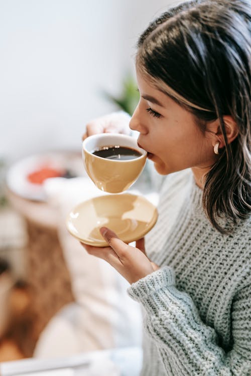 High angle side view of crop Asian female sipping aromatic brewed coffee in mug at home