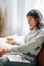 Side view of Asian female with mug of drink flipping page of magazine while reading with interest