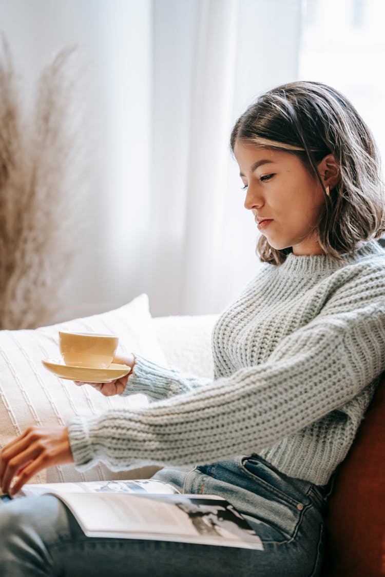 Serious Ethnic Woman With Cup Turning Page Of Magazine