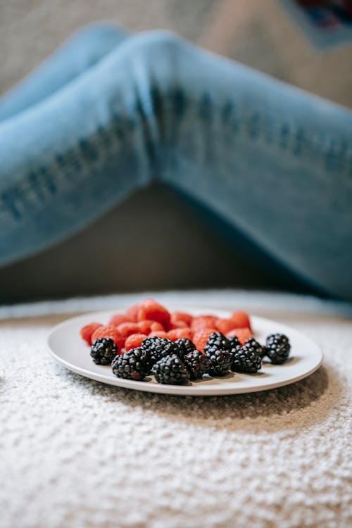 Free Crop anonymous female sitting on sofa with plate of yummy raspberries and blueberries at home Stock Photo