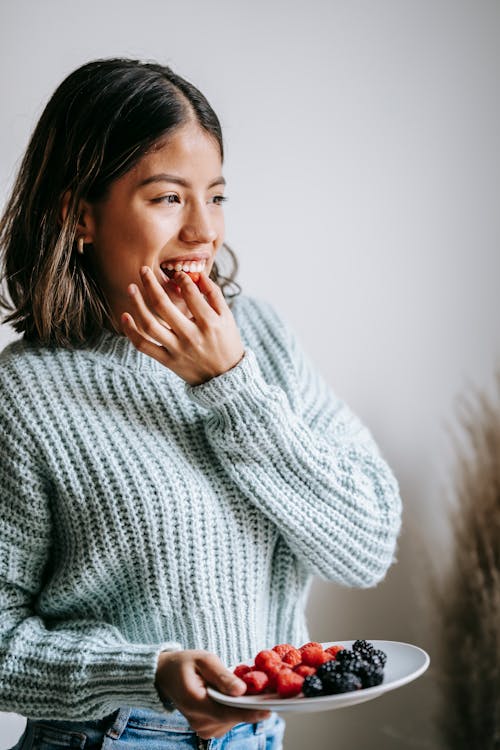 Positive Asian female in knitted sweater biting fresh healthy raspberry standing with plate of blueberries and looking away