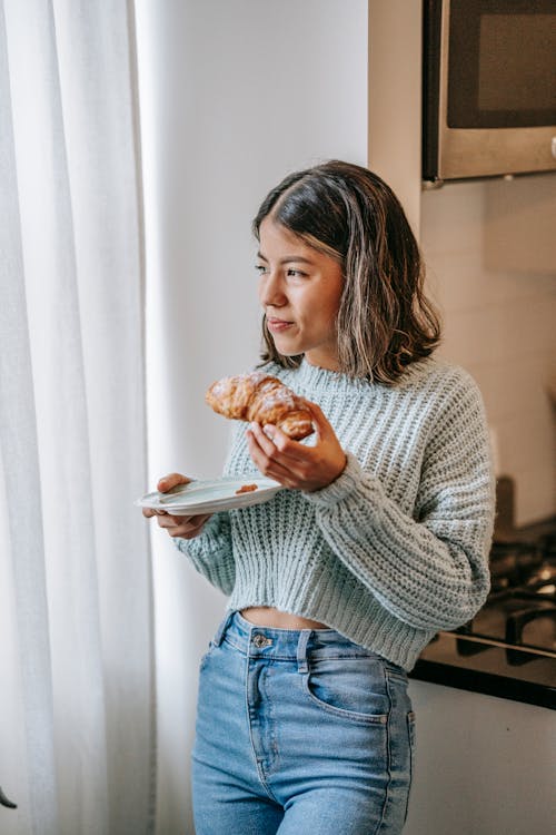 Smiling ethnic woman eating croissant in kitchen