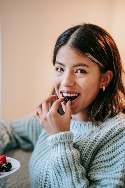 Happy Asian female in warm sweater enjoying tasty berry while eating healthy food at home