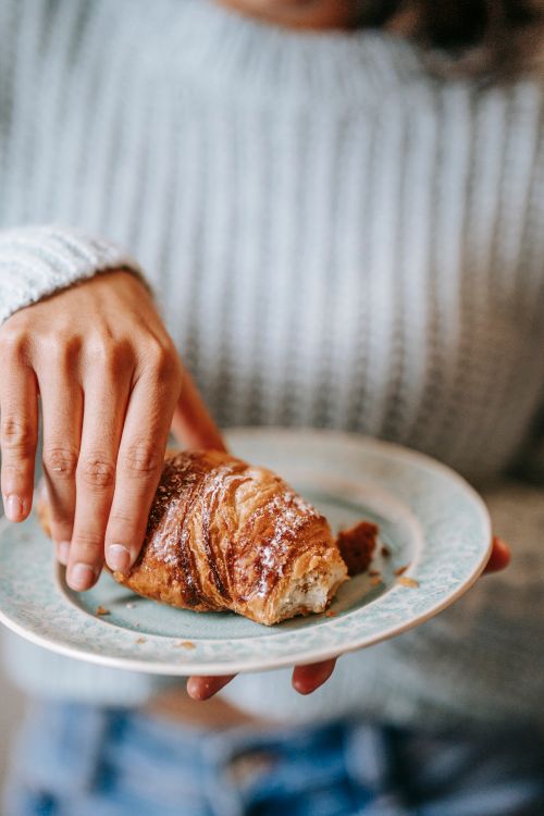 Free Crop unrecognizable lady in casual outfit with delicious croissant covered in white powder on plate in hands in light room Stock Photo