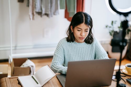 Free Young Latin American woman in casual outfit at table surfing on netbook near notepad with pen and cup with drink near ring light with phone near wardrobe and boxes Stock Photo