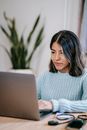 Confident young Hispanic woman in casual clothes working remotely on netbook at table in bright apartment with potted plant with green leaves near wall