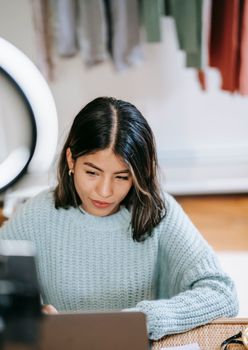 Confident young Latin American woman in casual clothes working remotely on laptop at table near ring light with phone near wardrobe with apparels in room