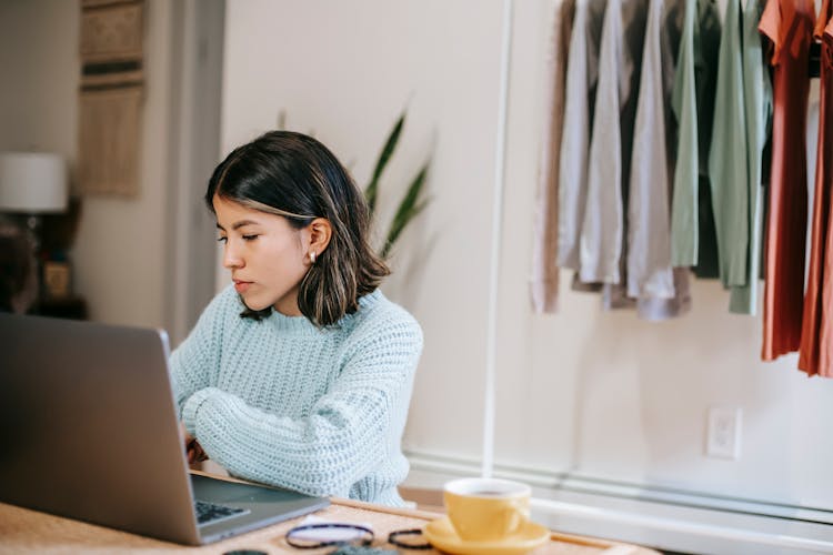 Serious Hispanic Lady Using Netbook Near Coffee Cup And Wardrobe