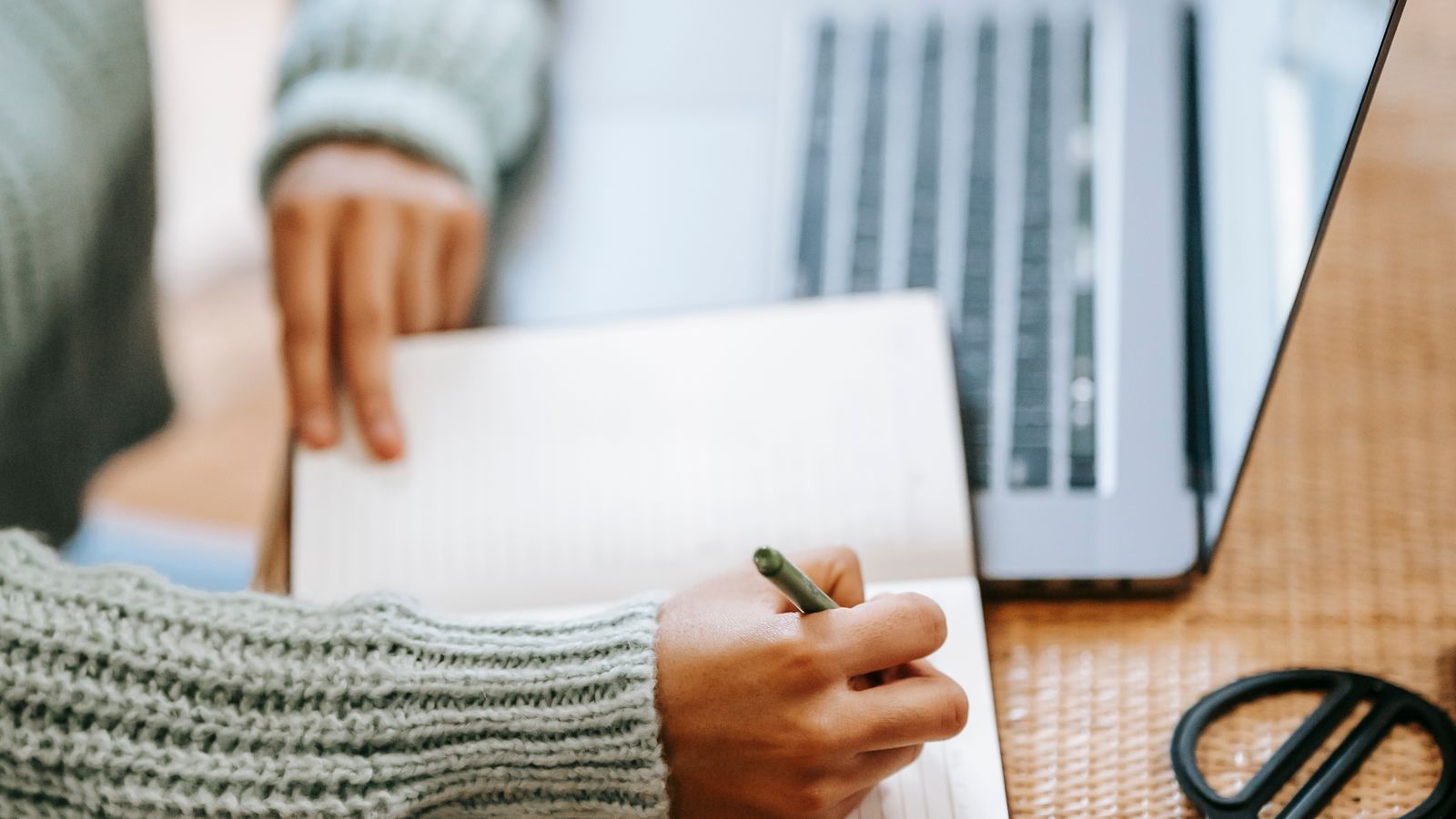A person writing in a notebook beside a laptop