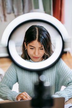 Confident young Hispanic woman in casual outfit at table browsing on netbook near ring light with cellphone near wardrobe with clothes in bright apartment
