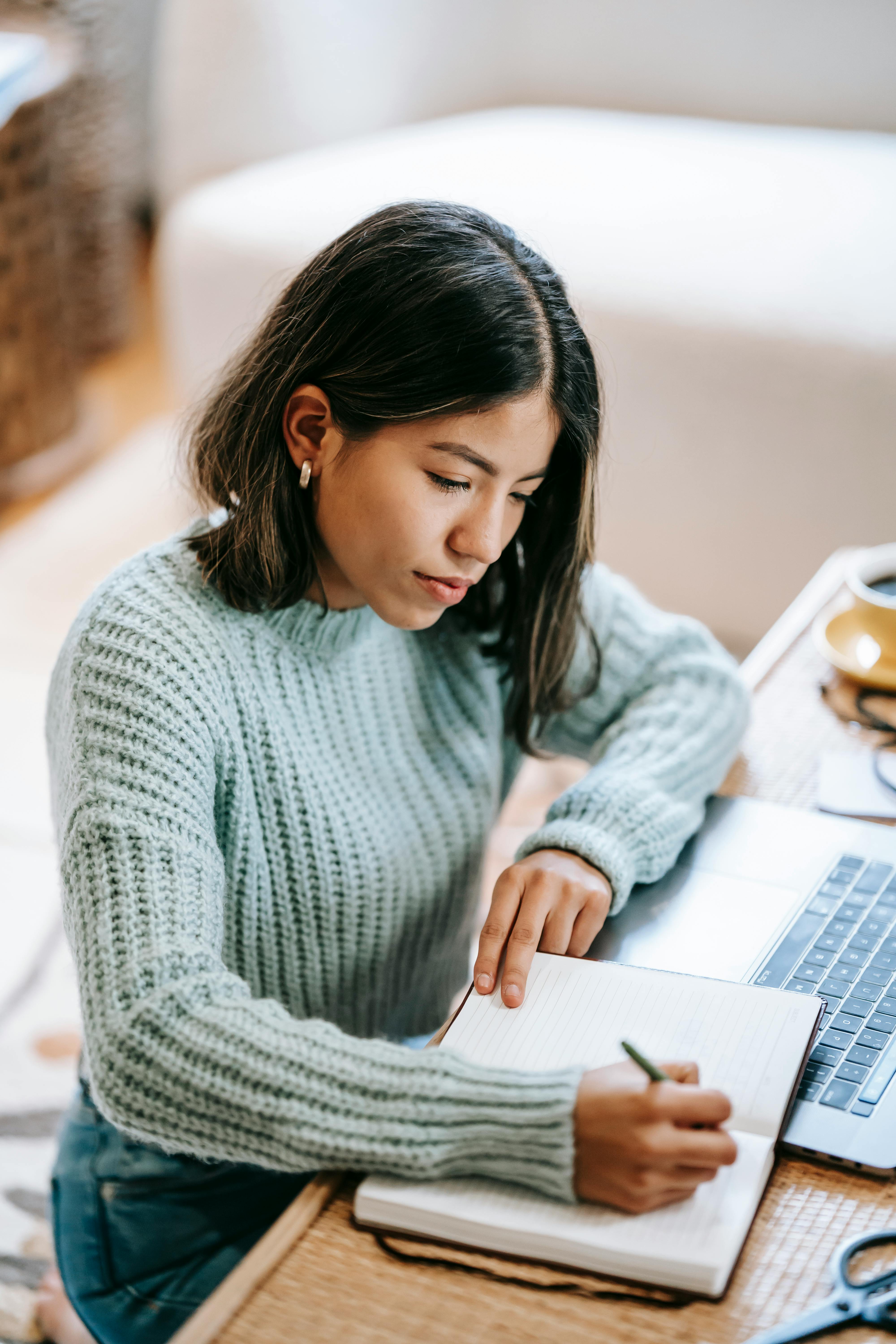 concentrated latin american lady writing information in notepad near netbook