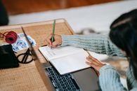High angle of anonymous ethnic female student in casual outfit taking notes with pen in notebook near laptop at table near scissors and ball of yarn on stamps at home