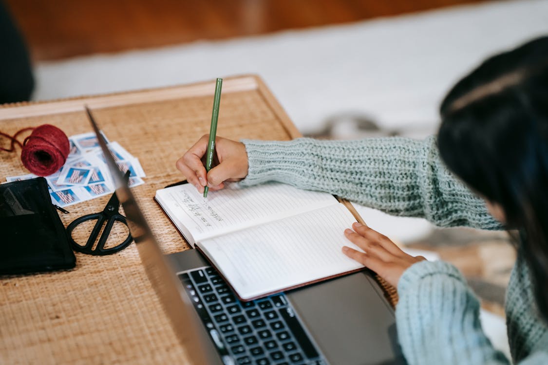 Free High angle of anonymous ethnic female student in casual outfit taking notes with pen in notebook near laptop at table near scissors and ball of yarn on stamps at home Stock Photo