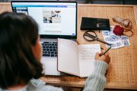 High angle of anonymous ethnic woman in casual clothes writing information in notepad at table near netbook and scissors near stamps and balls of thread in bright apartment