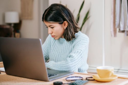 Free Serious young Latin American woman in casual clothes working remotely on netbook at table near mug with drink in light room with plant with green leaves and wardrobe with apparels Stock Photo