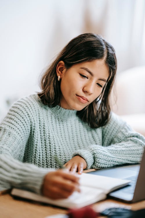 Free Young Hispanic female student in casual outfit using laptop while taking notes in notepad with pen at table in light apartment Stock Photo