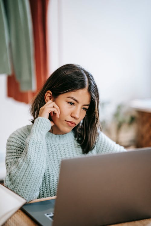 Youthful Latin American female in casual clothes sitting at table with laptop while looking at screen attentively at home