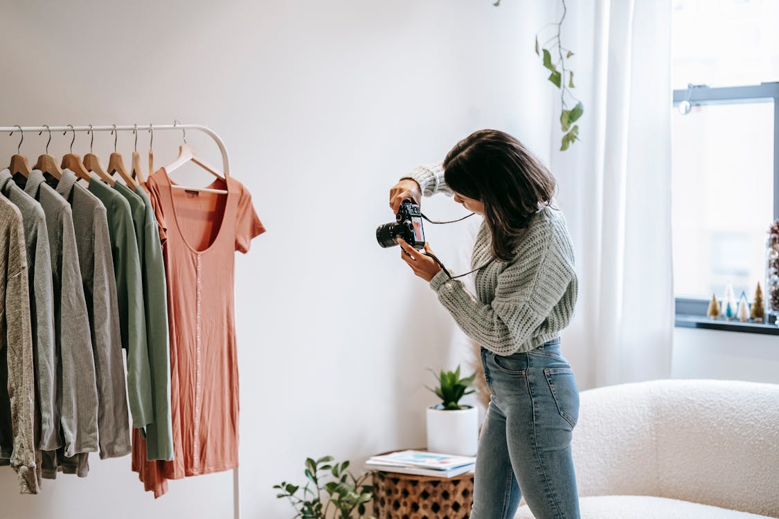 Free Side view of young female photographer in casual outfit standing in front of hanger and taking picture with camera of shirt in modern apartment Stock Photo