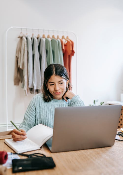 Happy young female freelancer taking notes and working on computer while sitting in front of clothing rack at wooden table in modern apartment