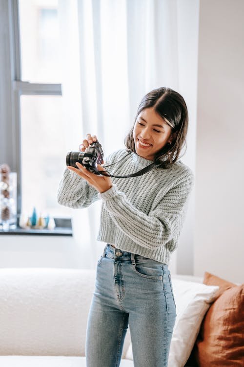 Positive young Latin American female photographer in casual clothes standing in light apartment while taking photos on professional photo camera near couch with pillows and window with curtains
