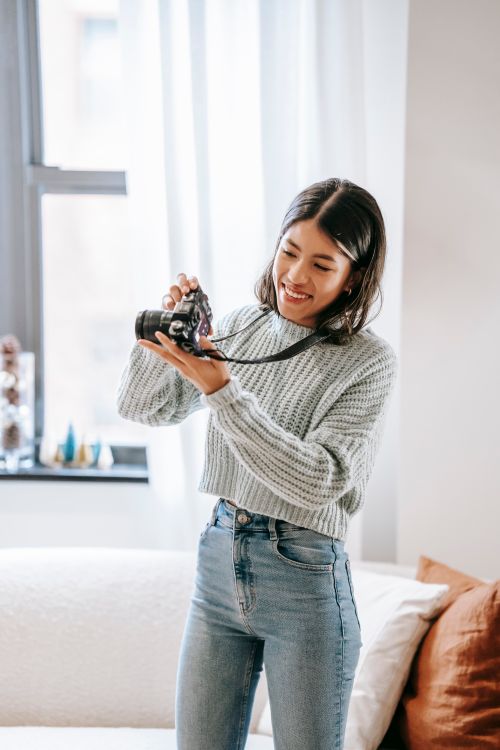 Free Positive young Latin American female photographer in casual clothes standing in light apartment while taking photos on professional photo camera near couch with pillows and window with curtains Stock Photo