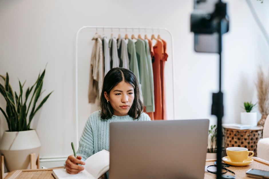 Serious young Latin American lady in casual clothes sitting at table while working remotely on laptop with notebook and pen near cup with drink and phone on stick in home