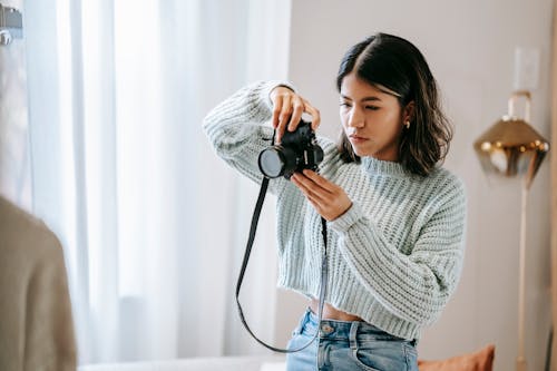 Serious young Hispanic woman in casual outfit taking photos on photo camera while standing at bright home near window with curtains