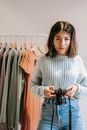 Latin American female in knitted sweater with professional photo device looking at camera against stand with clothes