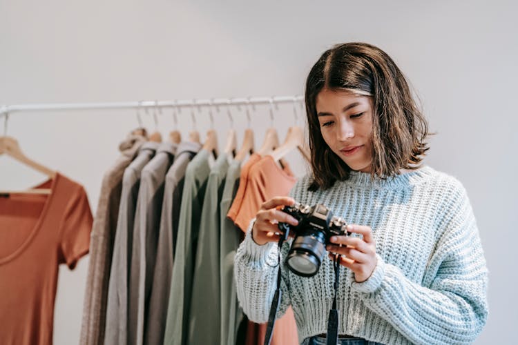 Ethnic Photographer With Digital Photo Camera Against Rack With Wear