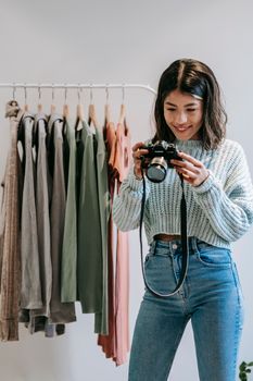 Young smiling Latin American woman watching professional photo camera against rack with assorted outfit indoors
