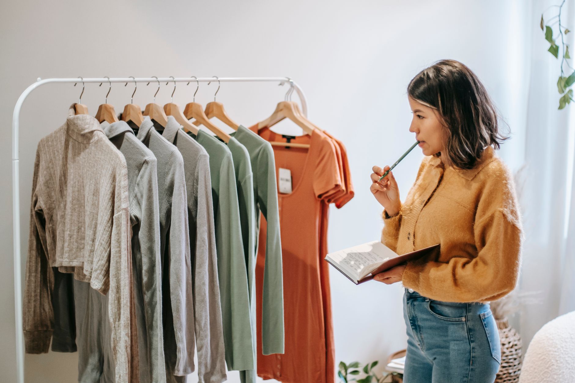 Side view of thoughtful Latin American female employee with diary biting pen while standing against assorted outfit at home