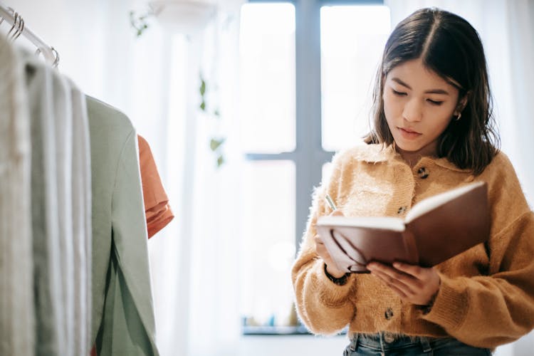 Ethnic Designer Writing In Journal Near Assorted Clothes At Home