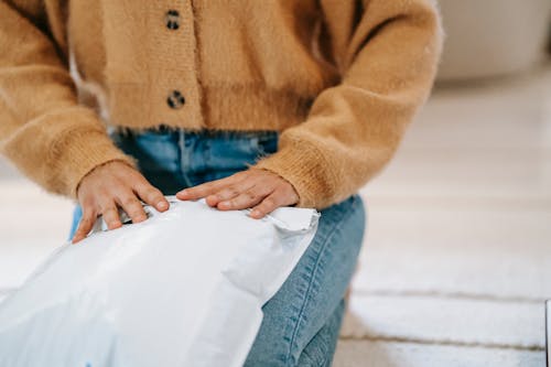 Crop faceless woman in casual clothes sitting on carpet while wrapping parcel bag for shipping in light room