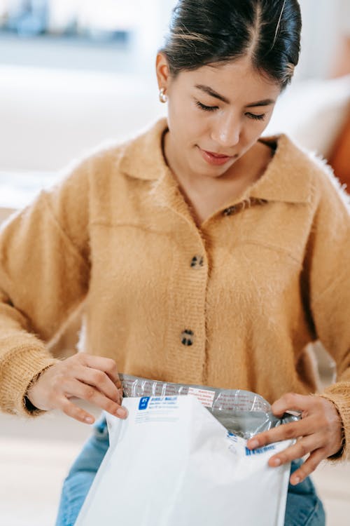 Confident young Latin American female in casual outfit sitting on carpet while unpacking shipping parcel bag in light apartment