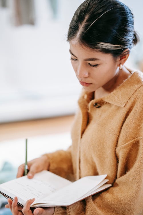 Free Side view of young Hispanic female in casual clothes with pen reading information from notebook in light room Stock Photo