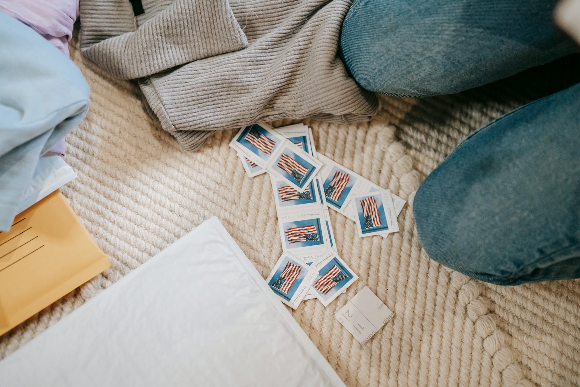 From above crop anonymous person in jeans sitting on carpet on floor near heaped US postage stamps and envelope