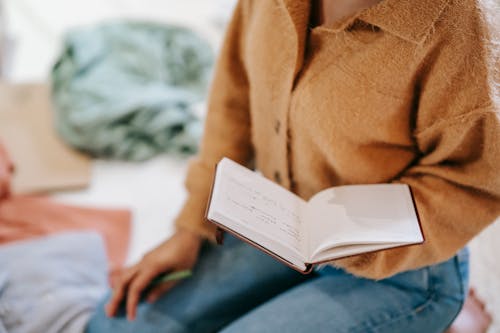 Crop anonymous female in casual clothes reading notes in notebook while sitting on floor in light room