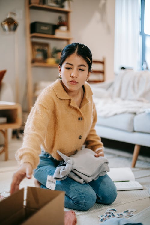 Focused young Asian female wearing casual outfit packing clothes into carton box while sitting on floor in modern living room