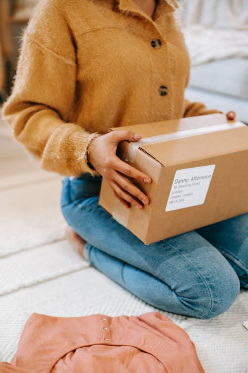 Crop anonymous female in casual outfit sitting on floor with carton parcel on laps in light room
