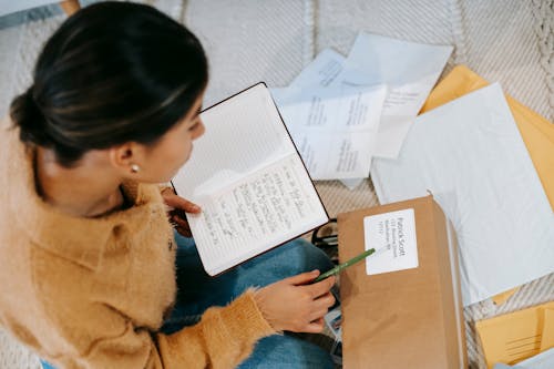 From above concentrated young female checking address details on parcel label while sitting on floor with opened diary