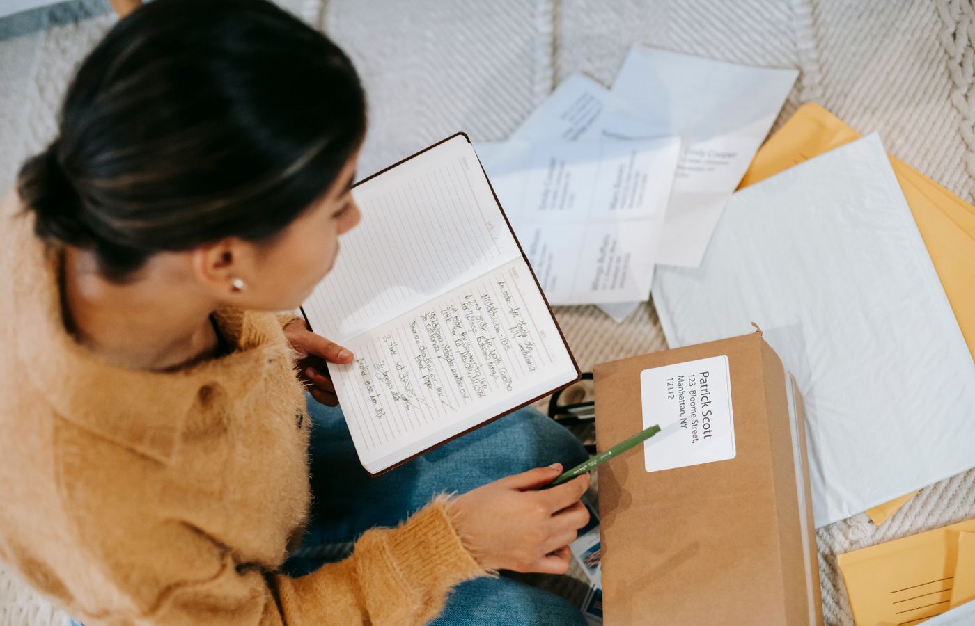 A young woman sitting on the floor as she organizes mail and packages, reading notes in a diary.