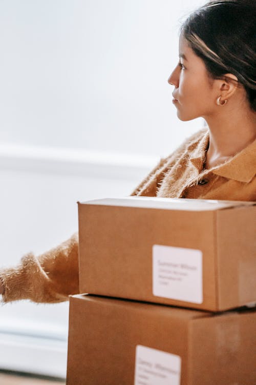 Free Crop concentrated Asian female in warm sweater standing with carton boxes in light post office and looking away thoughtfully Stock Photo