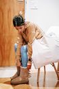 Focused young Asian female in jeans and warm sweater sitting on chair with big bag and putting on winter boots in light entrance hallway