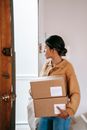 Focused young Asian female in casual clothes carrying cardboard parcels and big bag while leaving apartment