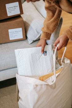 Crop unrecognizable female in warm sweater putting envelopes and parcels into big bag while standing near sofa with carton boxes
