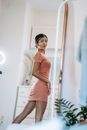 Beautiful fit Asian female wearing pink mini dress looking over shoulder in big mirror while standing in light bedroom