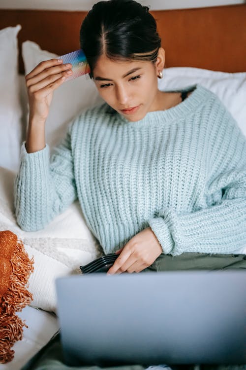 Thoughtful Asian woman with credit card using laptop on bed