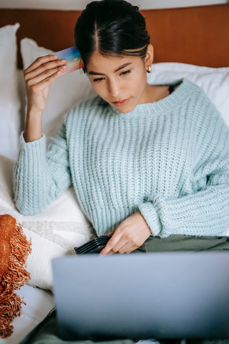 Thoughtful Asian Woman With Credit Card Using Laptop On Bed