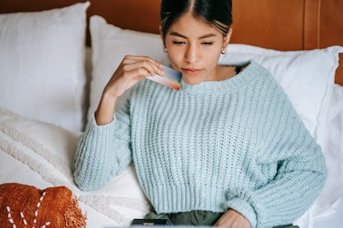 Crop concentrated Asian female in knitted blue sweater lying on cozy bed with credit card