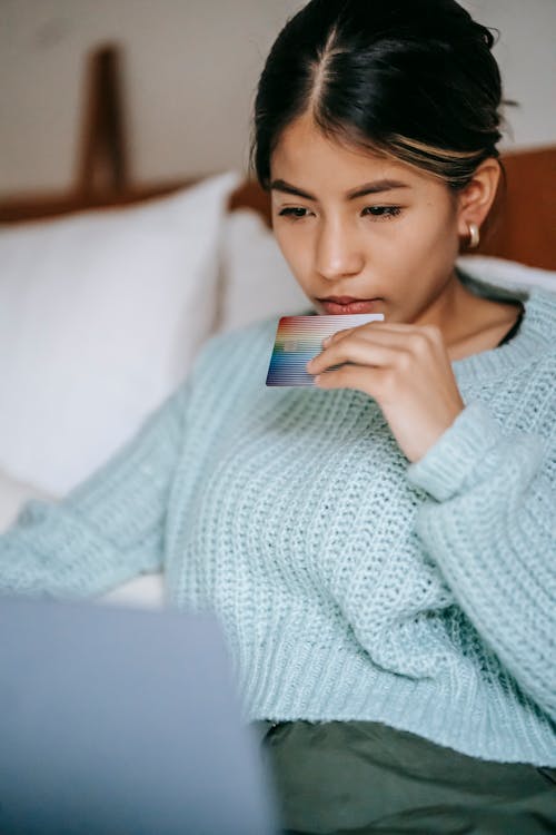 Crop concentrated Asian female wearing blue sweater making online payment while browsing netbook and lying on bed with credit card